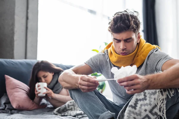 Selective focus of sick boyfriend looking at thermometer and holding napkin — Stock Photo