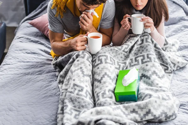 Cropped view of sick girlfriend and boyfriend sneezing and holding cups of tea — Stock Photo