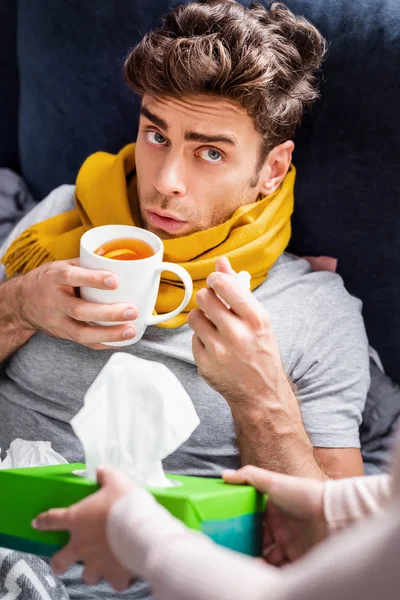 Cropped view of girlfriend giving box with napkins to sick boyfriend with cup of tea — Stock Photo