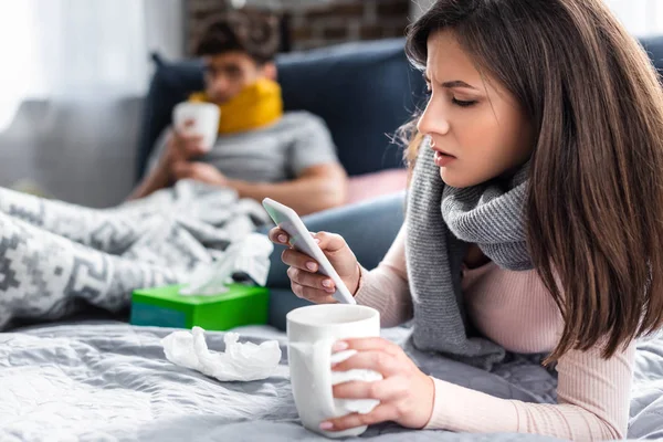 Selective focus of sick girlfriend holding cup of tea and using smartphone — Stock Photo