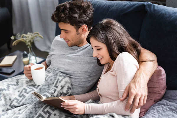Smiling girlfriend reading book and boyfriend holding cup of tea — Stock Photo