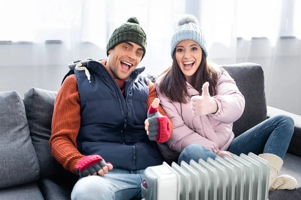 Smiling girlfriend and boyfriend in winter outfit showing thumbs up and warming up near heater — Stock Photo