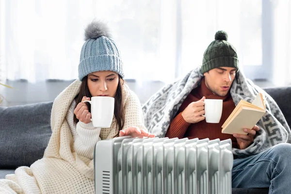 Attractive girlfriend with cup of tea and boyfriend reading book and warming up near heater — Stock Photo