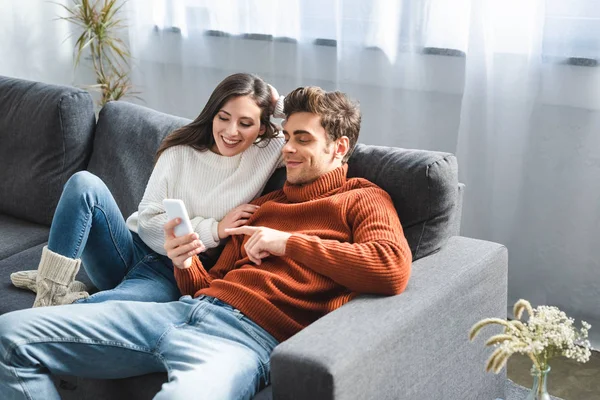 Novia sonriente y novio en suéteres mirando el teléfono inteligente - foto de stock