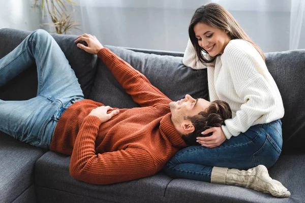 Namorado deitado de joelhos de namorada atraente em suéter e sorrindo no apartamento — Fotografia de Stock