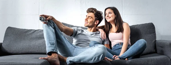 Tiro panorâmico de namorada atraente e namorado bonito com controle remoto sentado no sofá — Fotografia de Stock