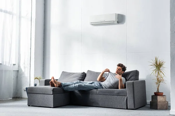 Handsome man in jeans looking at t-shirt and lying on sofa — Stock Photo