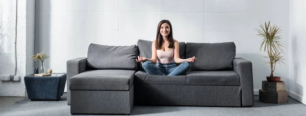 Prise de vue panoramique de femme attrayante et souriante en pose de lotus assis sur le canapé — Photo de stock