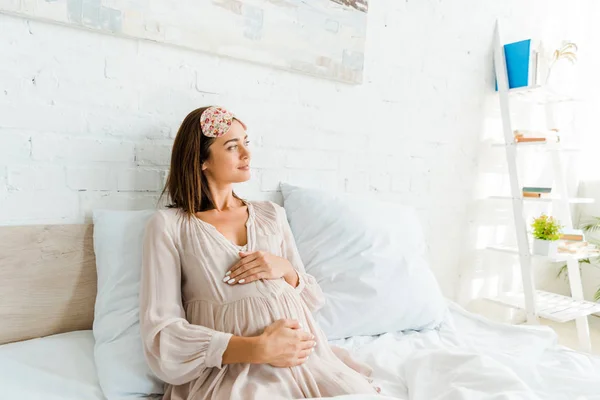 Schwangere mit Schlafmaske berührt ihren Bauch, während sie morgens im Bett liegt — Stockfoto