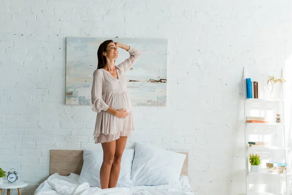 Hermosa mujer embarazada sonriente tocando su vientre mientras está de pie en la cama en casa - foto de stock