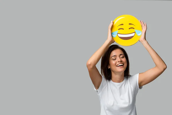 KYIV, UKRAINE - SEPTEMBER 10, 2019: cheerful woman holding yellow laughing emoji, isolated on grey — Stock Photo