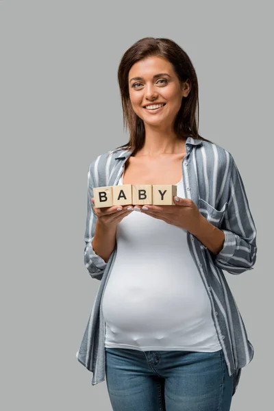 Красива усміхнена вагітна жінка тримає кубики абетки з дитячим знаком, ізольована на сірому — стокове фото