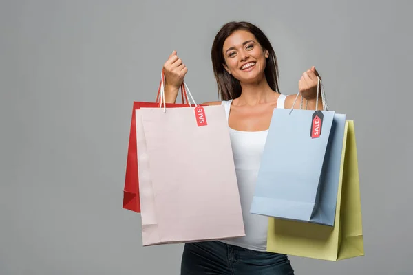 Mulher sorrindo segurando sacos de compras com sinais de venda, isolado em cinza — Fotografia de Stock