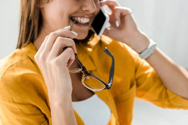 Recortado vista de sonriente freelancer sosteniendo gafas y hablando en el teléfono inteligente - foto de stock