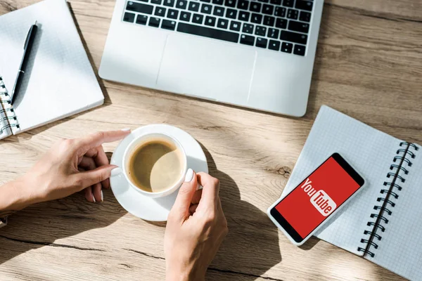 KYIV, UKRAINE - SEPTEMBER 10, 2019: cropped view of freelancer with cup of coffee, laptop and smartphone with youtube app — Stock Photo