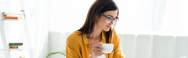 Bela freelancer feminino em óculos segurando xícara de café no escritório em casa — Fotografia de Stock