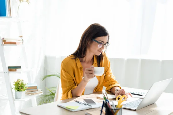 Belo freelancer trabalhando no laptop com xícara de café no escritório em casa — Fotografia de Stock