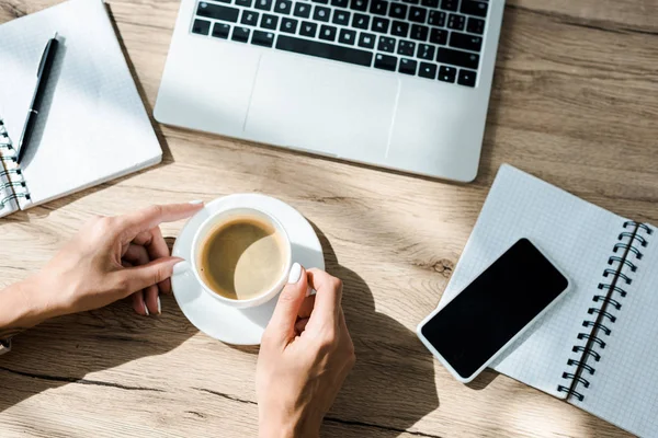 Cropped view of freelancer with coffee, notebooks, laptop and smartphone on table — Stock Photo