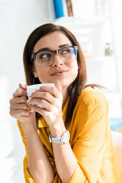 Attractive pensive freelancer in eyeglasses holding cup of coffee in home office — Stock Photo