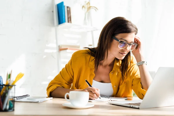 Freelancer feminino atraente trabalhando no laptop e escrevendo em home office — Fotografia de Stock