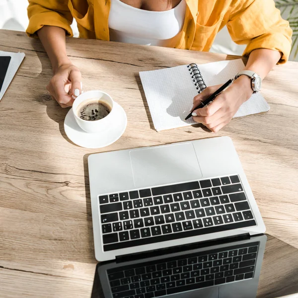 Cropped view of freelancer working with laptop, writing in notebook and holding cup of coffee in home office — Stock Photo