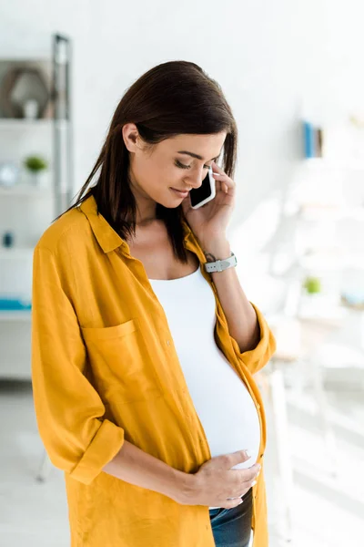 Schöne schwangere Freiberuflerin im gelben Hemd, die im Homeoffice auf dem Smartphone spricht — Stockfoto