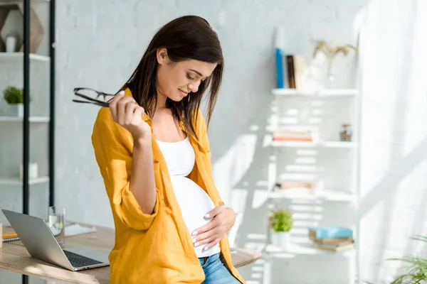 Счастливый беременный фрилансер трогает животик, сидя в домашнем офисе — стоковое фото