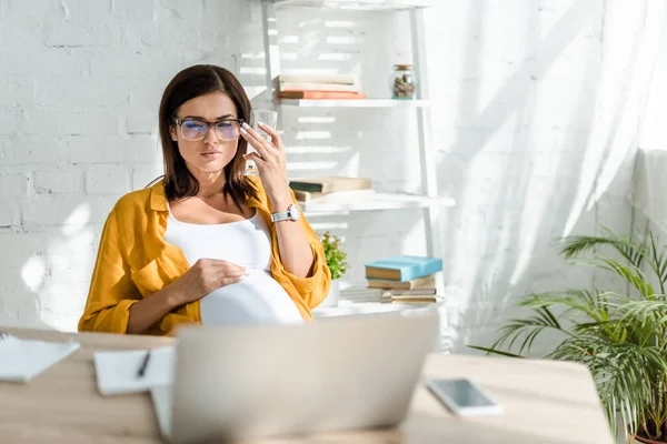 Pregnant freelancer holding glass of water in office and looking at home office — Stock Photo