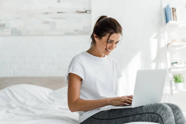 Mujer joven sonriente usando el ordenador portátil en la cama por la mañana - foto de stock