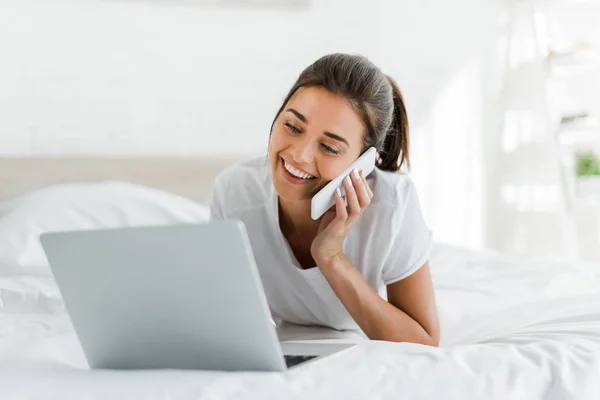 Chica sonriente atractiva hablando en el teléfono inteligente y el uso de ordenador portátil en la cama por la mañana - foto de stock