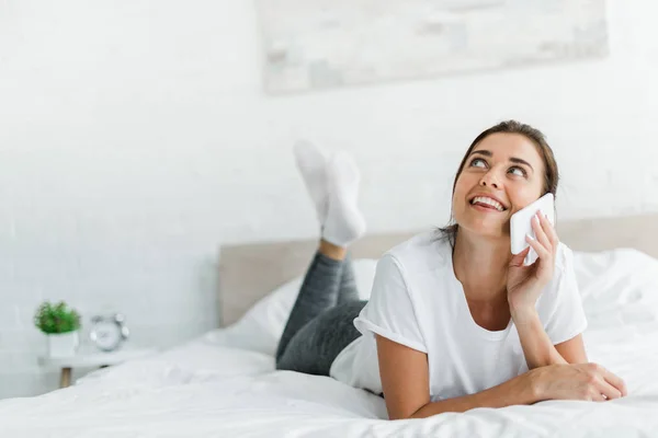 Привлекательная веселая девушка разговаривает по смартфону, лежа на кровати — стоковое фото