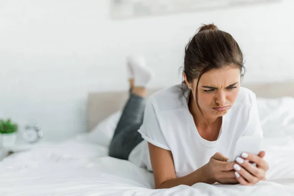 Attractive confused girl using smartphone while lying on bed in the morning — Stock Photo