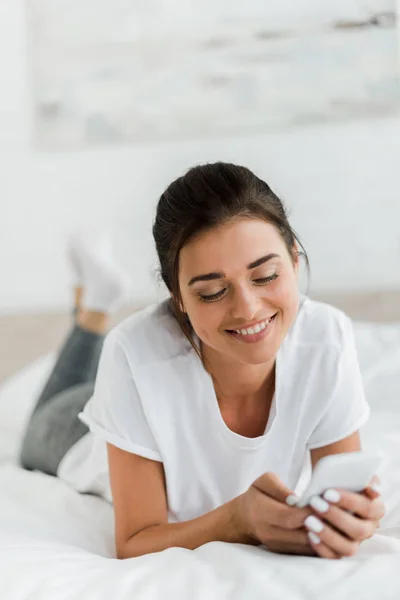Happy girl using smartphone while lying on bed in the morning — Stock Photo