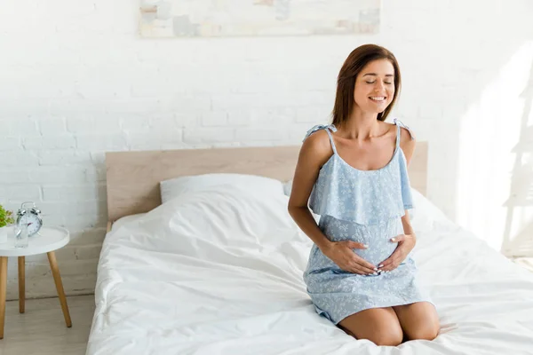 Улыбающаяся беременная женщина в синем платье трогает живот в спальне — стоковое фото
