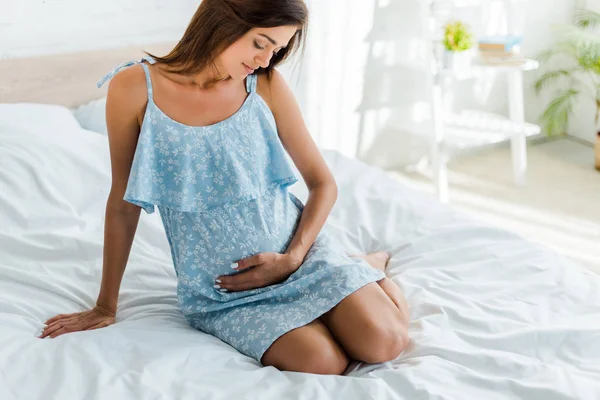 Beautiful pregnant woman in blue dress touching her belly in bedroom — Stock Photo