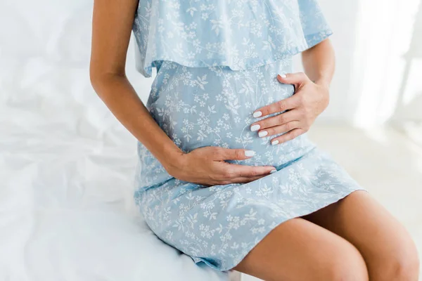 Cropped view of pregnant woman in blue dress touching her belly in bedroom — Stock Photo