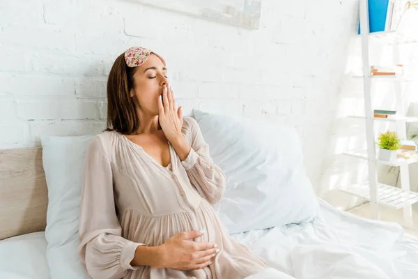 Mujer embarazada con máscara de dormir bostezando en la cama por la mañana - foto de stock