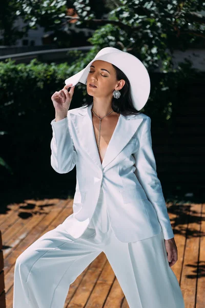 Mulher atraente em terno branco e chapéu posando com olhos fechados fora — Fotografia de Stock