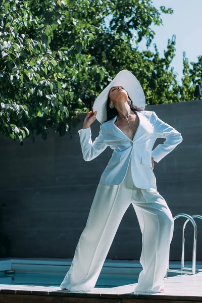 Jolie femme en costume blanc et chapeau posant avec les yeux fermés à l'extérieur — Photo de stock