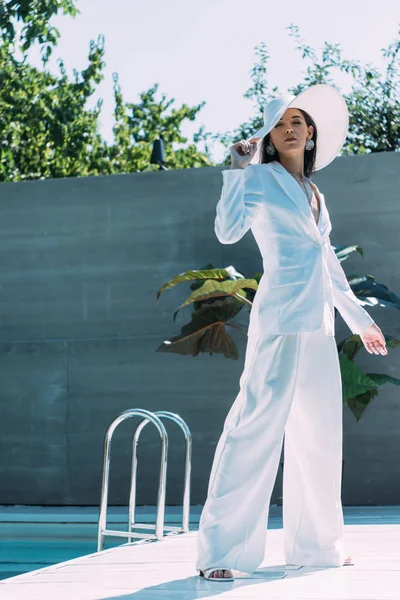 Atractiva mujer en traje blanco y sombrero posando y mirando a la cámara exterior - foto de stock
