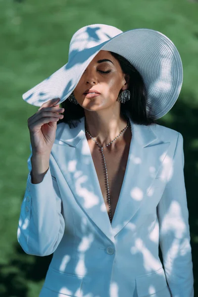 Attractive woman in white suit and hat posing with closed eyes outside — Stock Photo