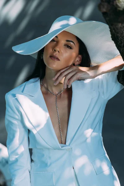 Attrayant femme en costume blanc et chapeau posant et regardant la caméra à l'extérieur — Photo de stock