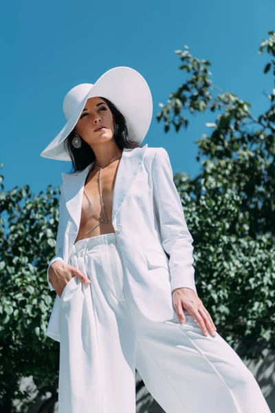Visão de ângulo baixo de mulher atraente em terno branco e chapéu posando fora — Fotografia de Stock