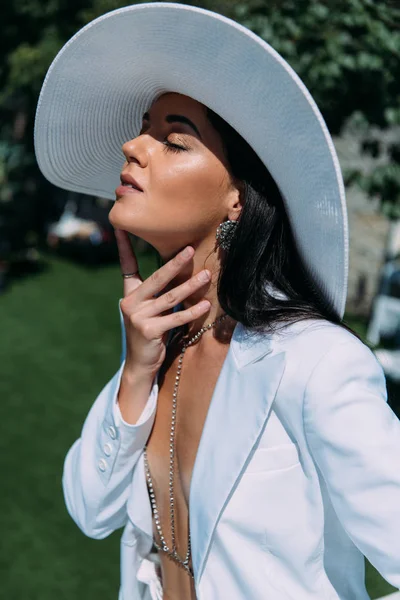Привлекательная женщина в белом костюме и шляпе позирует с закрытыми глазами снаружи — стоковое фото
