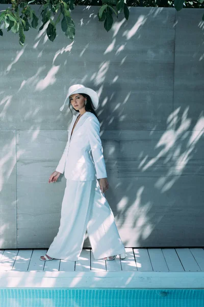 Atractiva mujer en traje blanco y sombrero caminando y mirando a la cámara exterior - foto de stock