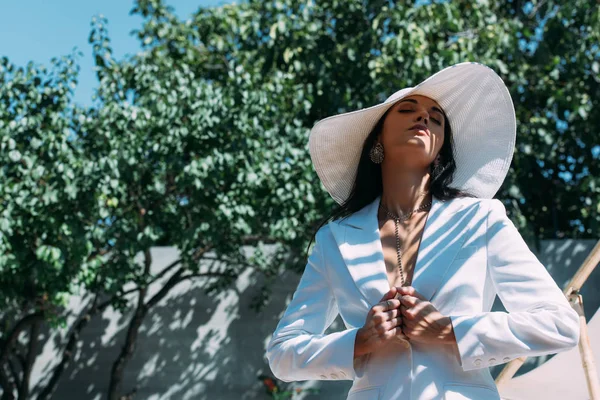 Vista de ángulo bajo de mujer atractiva en traje blanco y sombrero posando fuera — Stock Photo