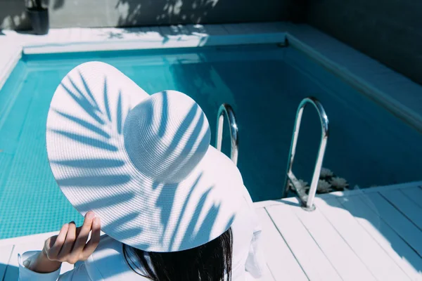 Vue arrière de la femme en chapeau blanc assis près de la piscine à l'extérieur — Photo de stock