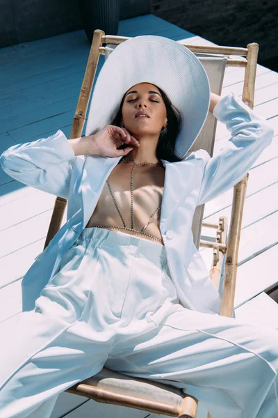 Atractiva mujer en traje blanco y sombrero sentado om silla de cubierta con los ojos cerrados fuera — Stock Photo