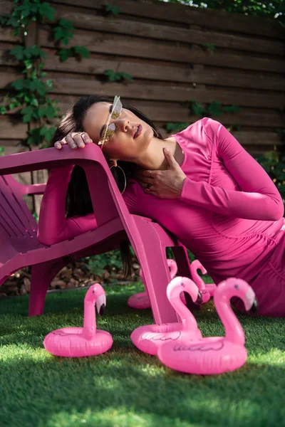 Attrayant femme en robe et lunettes de soleil posant et assis sur l'herbe à l'extérieur — Photo de stock