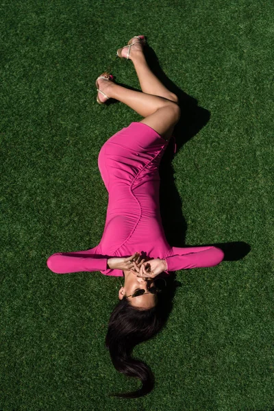 Vista superior da mulher atraente em vestido e óculos de sol posando e deitado na grama fora — Fotografia de Stock